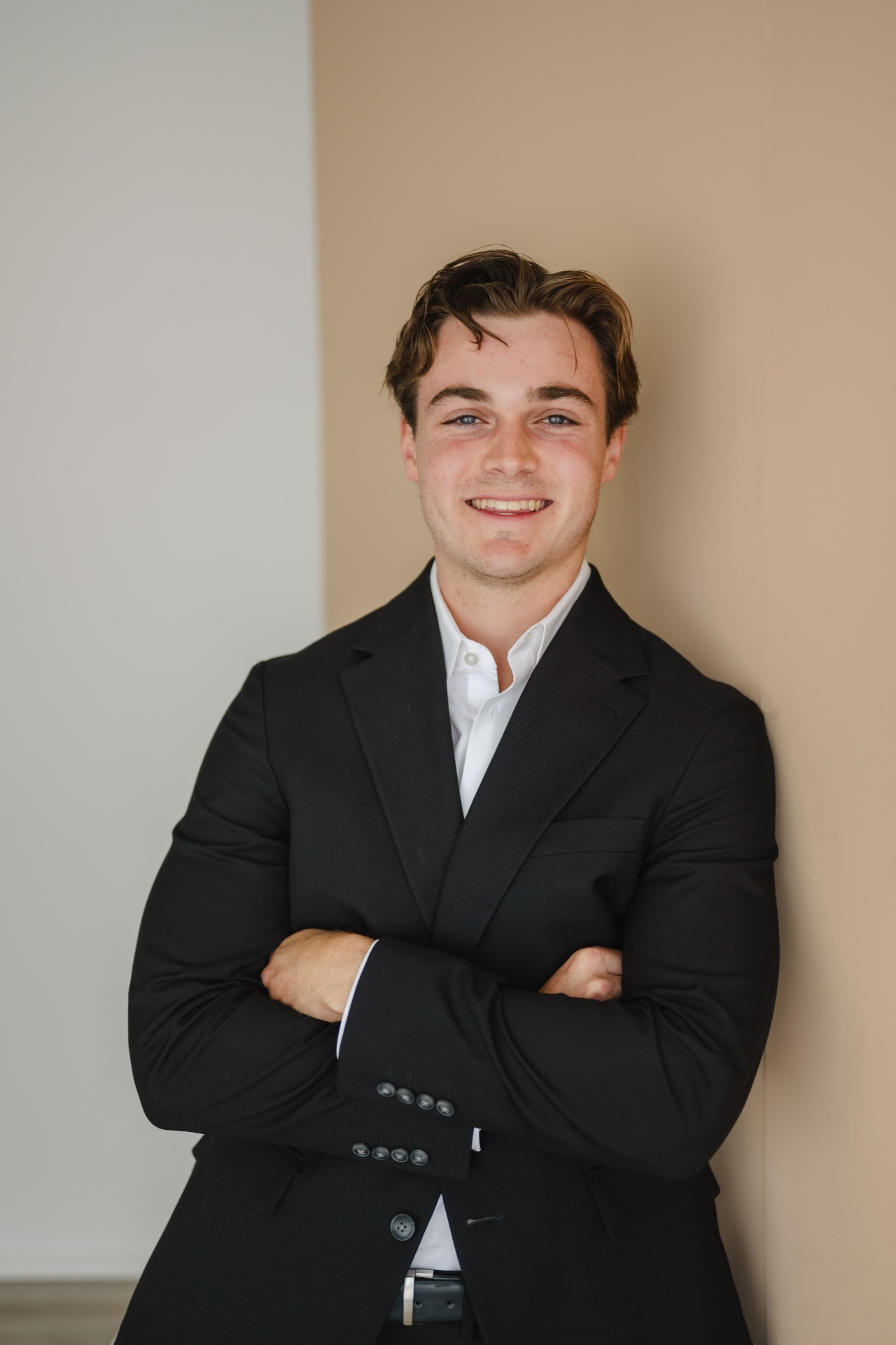 Hunter Smith in a black suit, arms crossed, leaning against a warm neutral wall